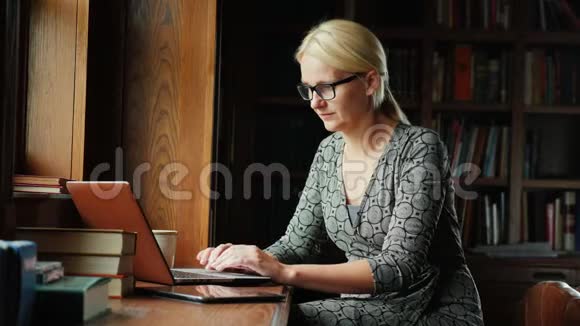 戴眼镜的女商人在图书馆或豪华办公室里用笔记本电脑工作在后台书架上放着书视频的预览图