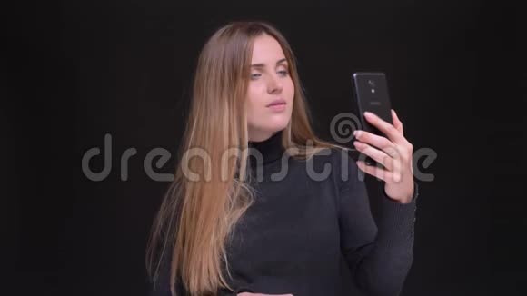 年轻的白种人长发金发金发女孩在黑色背景下使用智能手机制作自拍照片的肖像视频的预览图