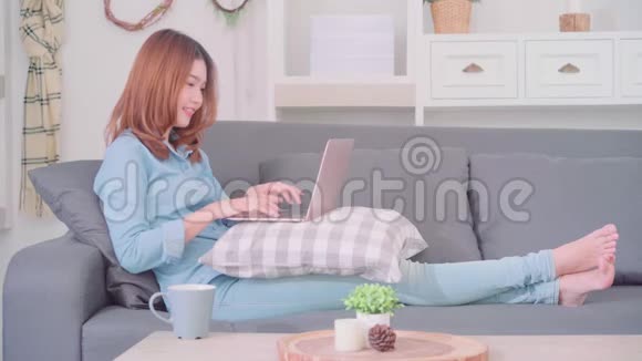 在家里的客厅里放松时亚洲女人在沙发上躺着用电脑或笔记本电脑拿着一杯热咖啡或茶视频的预览图