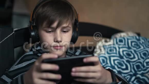 金发男孩戴着黑色耳机听音乐在家里用黑色手机玩网络游戏视频的预览图