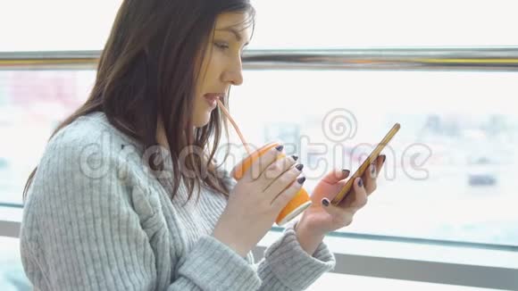一个穿灰色毛衣的年轻女子在窗口的咖啡馆里拿着手机喝果汁视频的预览图