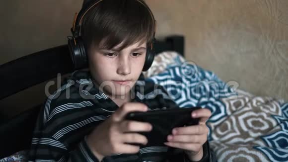一个快乐的少年男孩躺在床上拿着智能手机和耳机在家里听音乐儿童休息视频的预览图