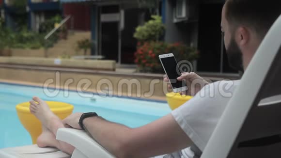 成年男子正在游泳池附近滚动他的智能手机屏幕视频的预览图