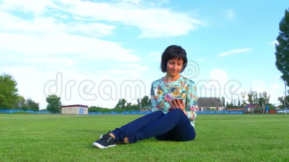 坐在体育场草坪上的年轻女子在多云的蓝天背景下使用智能手机视频的预览图