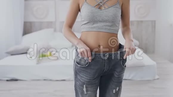 年轻的女人展示她的减肥和穿她的旧牛仔裤在家里在轻卧室穿太大牛仔裤的苗条女孩视频的预览图