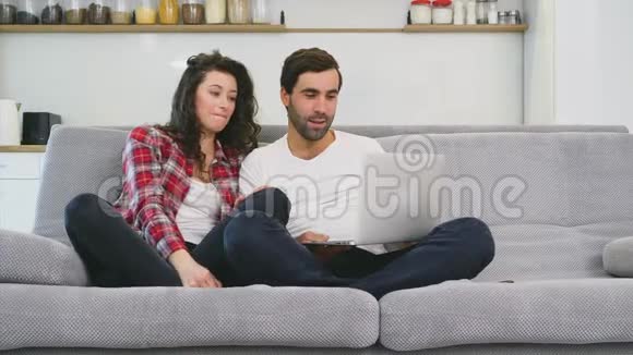 幸福的男人和幸福的男人在家里一起坐在沙发上微笑着的年轻夫妇用电脑享受早晨的咖啡视频的预览图