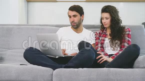 幸福的男人和幸福的男人在家里一起坐在沙发上微笑着的年轻夫妇用电脑享受早晨的咖啡视频的预览图
