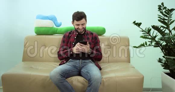 充满魅力的男人坐在沙发上看着他的手机他微笑着快乐着4k视频的预览图