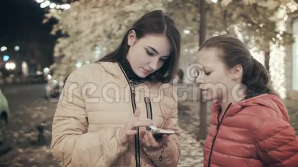 一个女人晚上和她的女儿一起在城市街道上使用智能手机视频的预览图