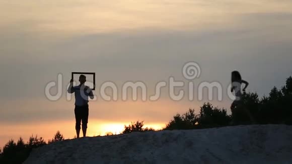 在夕阳的背景下一对滑稽有趣的浪漫情侣抱着空框接吻视频的预览图