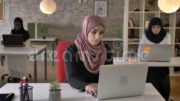 三个戴头巾的年轻穆斯林妇女坐在现代办公室的笔记本电脑上打字戴头巾的黑人妇女工作视频的预览图