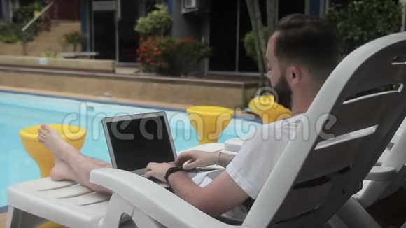 年轻人正拿着笔记本电脑坐在游泳池旁的休息室里视频的预览图