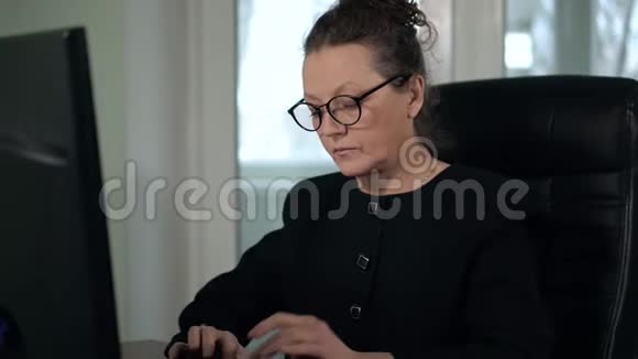 成熟的女人戴着眼镜穿着黑色西装坐在窗前的办公室里用电脑打字视频的预览图