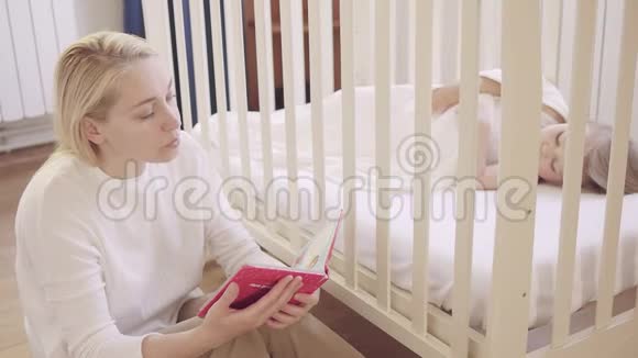 一个成年女人坐在婴儿床上睡觉的小婴儿旁边看书视频的预览图