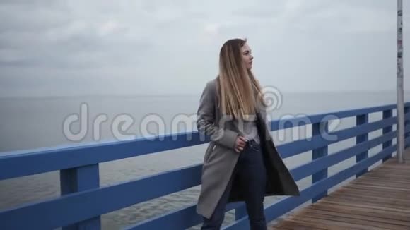 年轻快乐的女人金发穿着灰色外套和牛仔裤沿着海滨长廊散步视频的预览图