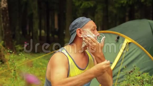 一个男人坐在一个旅游帐篷附近剃掉了脸上的头发视频的预览图