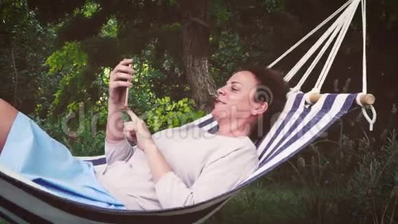 美丽的女孩躺在吊床上正在玩她的智能手机视频的预览图