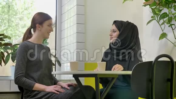 两个年轻漂亮的女人坐在咖啡馆里其中一个是戴着头巾的漂亮穆斯林女人谈笑风生视频的预览图