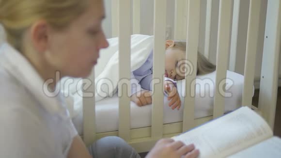 一个成年女人坐在婴儿床上睡觉的小婴儿旁边看书视频的预览图