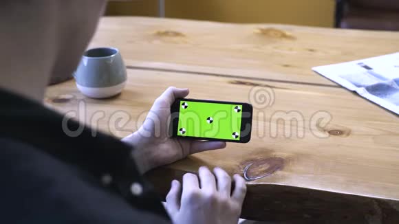 坐在木桌旁看着手机的彩色钥匙绿色屏幕一个穿着黑色衬衫的年轻人的肩膀上的景色视频的预览图