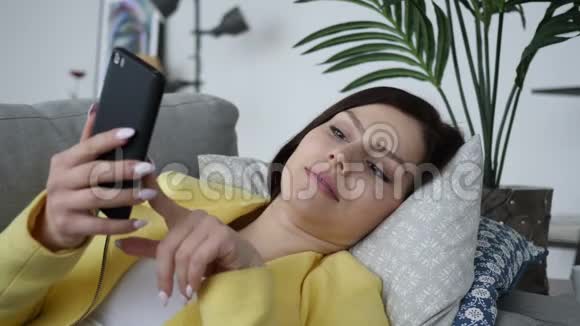 年轻女子在家里躺在沙发上忙着用智能手机视频的预览图