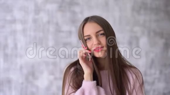 穿着粉色衬衫的快乐美女在智能手机前聊天她微笑着看着灰色背景下的相机视频的预览图