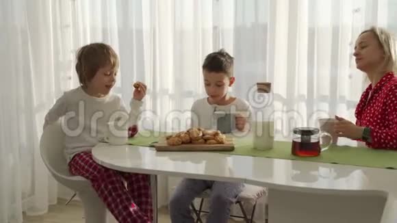 两个男孩在现代厨房吃早餐男孩吃带牛奶的饼干厨房里的白色桌子母亲和两个儿子视频的预览图