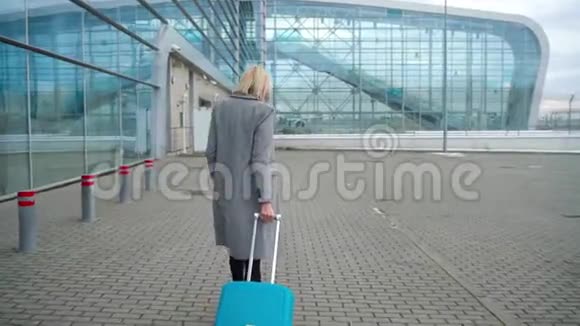 金发女孩在机场候机楼附近滚动一个手提箱从后面看视频的预览图