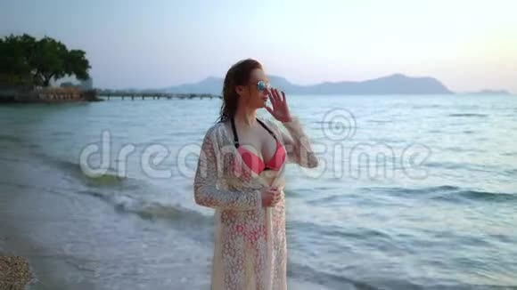 一个可爱的女人带着雀斑穿着粉红色的泳衣戴着太阳镜穿着白色的连衣裙在一个有棕榈树的白色沙滩上视频的预览图