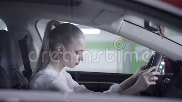 穿着白色衬衫的漂亮女人坐在车里拿着方向盘在车库旁边视频的预览图