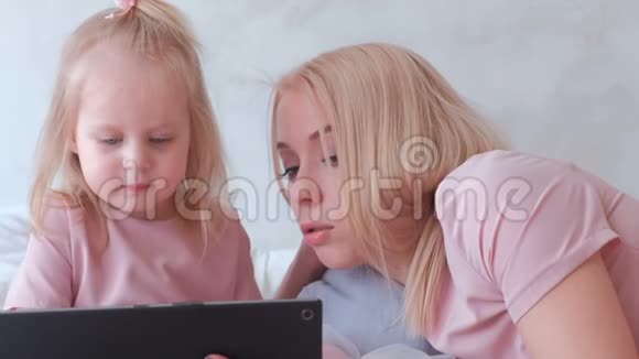 年轻迷人的金发女人用躺在床上的平板电脑教她穿着粉红色裙子的迷人小女儿特写视频的预览图