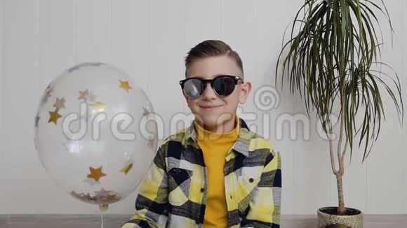 一个戴太阳镜的英俊小男孩坐在地板上背景是白色的墙手里拿着一个气球视频的预览图
