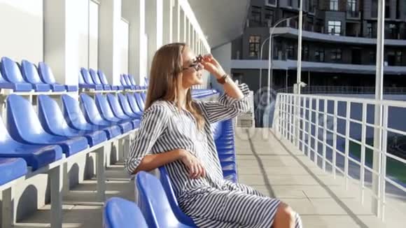 4K镜头戴太阳镜的年轻美女在体育场空座位上放松视频的预览图