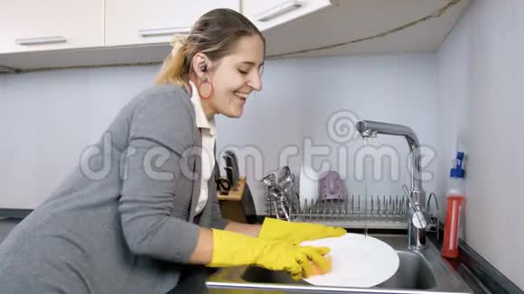 在厨房洗碗的时候带着耳机听音乐的快乐微笑女人的4k镜头视频的预览图