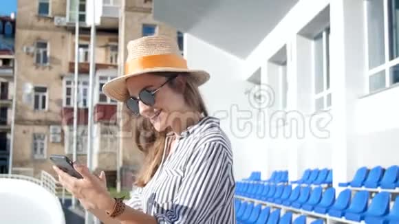 阳光明媚的一天快乐的年轻女子戴着太阳镜和帽子在街上使用无线电话视频的预览图