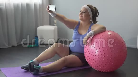 自信快乐的肥胖女性在家庭锻炼时在智能手机上自拍视频的预览图