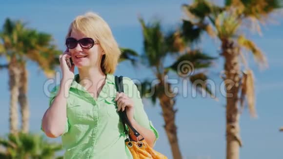戴太阳镜的女游客在天空和棕榈树的背景下打电话视频的预览图