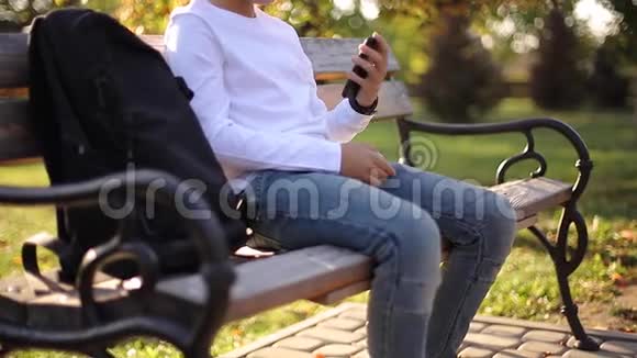 穿着白色运动衫背着黑色背包坐在公园里的长凳上和电话旁的人说话视频的预览图