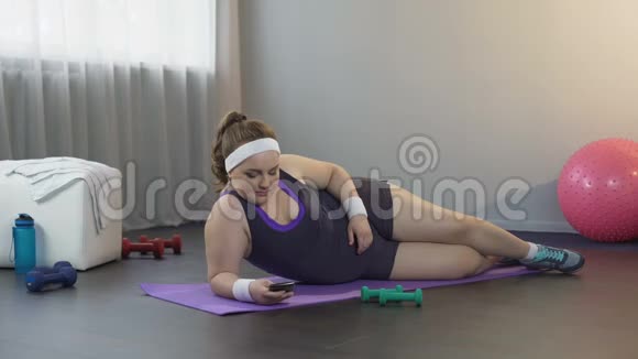 懒惰的胖女孩躺在垫子上在社交媒体上滚动小工具而不是锻炼视频的预览图