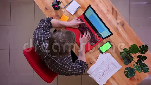 一位年轻的高加索商人在办公室室内的办公桌上用手机在笔记本电脑上打字的特写镜头视频的预览图