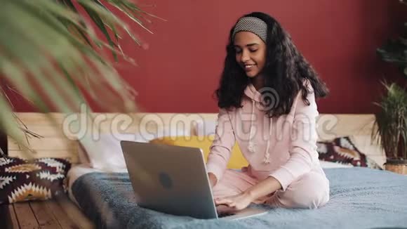 漂亮的女孩穿着粉红色睡衣家里坐在床上用笔记本电脑年轻女子微笑着视频的预览图