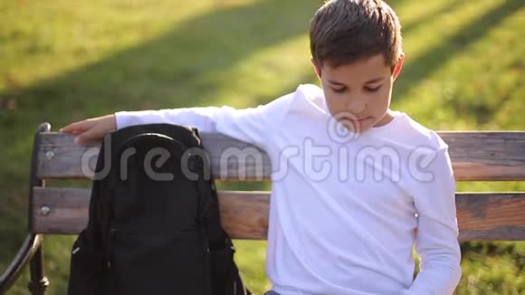 穿着白色运动衫背着黑色背包坐在公园里的长凳上和电话旁的人说话视频的预览图