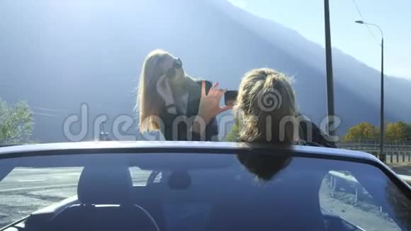 两个漂亮的年轻时尚女孩在红色敞篷车里的智能手机上拍照假期有趣的照片拍摄视频的预览图