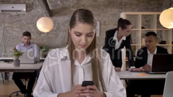 年轻漂亮的女商人正在办公室里用智能手机打字她的同事正在与科技建立联系视频的预览图
