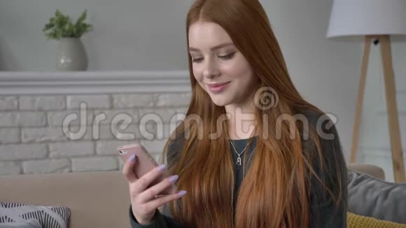 年轻漂亮的红发少女使用智能手机微笑发短信滚动滑动家庭舒适视频的预览图