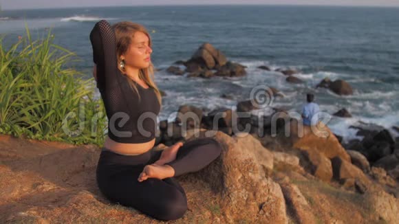 穿着运动服的女士坐在悬崖上在海边做瑜伽姿势视频的预览图