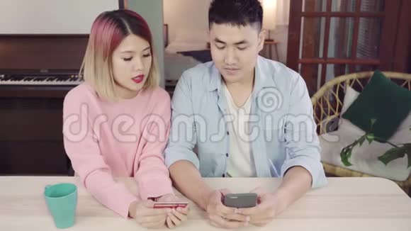 年轻的亚洲家庭夫妇使用智能手机讨论新闻或做网上购物一起坐在办公桌上的家庭周末视频的预览图