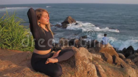 穿着运动服的女士坐在悬崖上在海边做瑜伽姿势视频的预览图