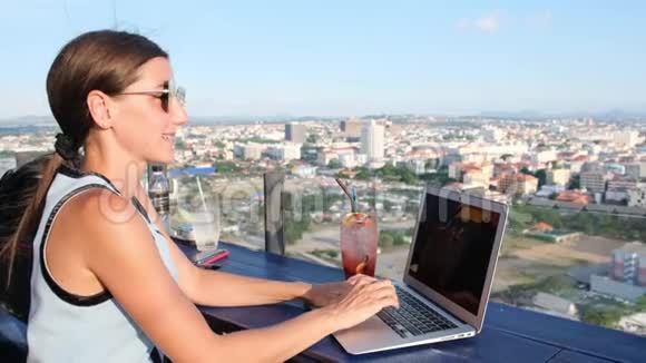 女性在一栋高楼屋顶的一家咖啡馆里的笔记本电脑上工作她可以看到城市的美丽全景视频的预览图