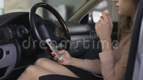 女办公室职员坐在车里吃三明治在智能手机上滚动视频的预览图